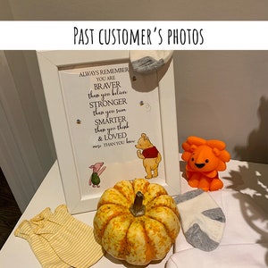 May include: A framed Winnie the Pooh quote that reads "Always remember you are braver than you believe, stronger than you seem, smarter than you think & loved more than you know."  The frame is on a white table with a small pumpkin, a stuffed lion, and two pairs of baby socks.