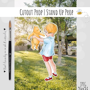 May include: A cutout prop of a boy holding a teddy bear. The boy is wearing a blue shirt, red shorts, and red shoes. The teddy bear is brown and has a yellow belly. The background is a blurry image of a park with green grass and trees.