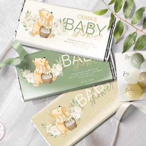 May include: Three green and white chocolate bar wrappers with a Winnie the Pooh character holding a honey pot. The wrappers have the text "Olivia's Baby Shower" and "Baby Shower" on them. The wrappers are decorated with white flowers and green leaves.