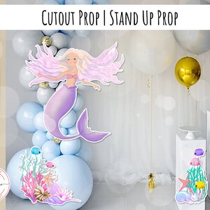 May include: A cutout prop of a mermaid with long purple hair and a tail. The mermaid is standing in front of a backdrop of blue and white balloons. The prop is 24 inches by 36 inches and is available as a JPG download.