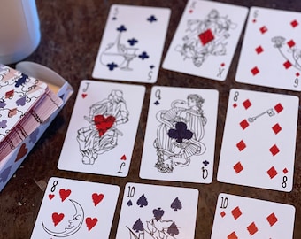 May include: A spread of playing cards on a wooden table. The cards feature a variety of suits, including hearts, diamonds, clubs, and spades. The cards are decorated with illustrations of various objects, including a crescent moon, a ship, and a book.