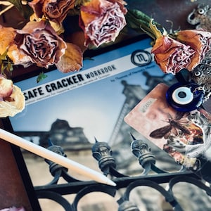 Może przedstawiać: Niebieski i biały amulet w kształcie złego oka wisi na srebrnym łańcuszku. Amulet leży na karcie tarota z nadrukowanym słowem "DEATH". Karta leży na książce zatytułowanej "SAFE CRACKER WORKBOOK: nation exercises for tapping into the secrets of Scorpio" autorstwa Thomasa of Hermit's Maror.