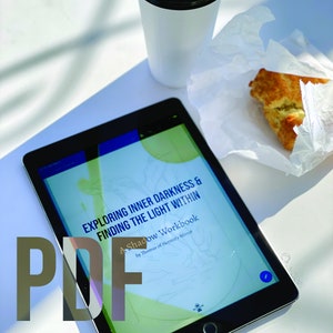 May include: A digital tablet displaying a PDF document titled "Exploring Inner Darkness & Finding the Light Within: A Shadow Workbook by Thomas of Hermit's Mirror". The tablet is on a white surface with a coffee cup and a pastry.