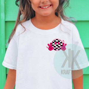 Puede incluir: Camiseta blanca con una cara sonriente a cuadros en negro y rosa con signos de paz. El gr&aacute;fico tiene dos estrellas rosas arriba.