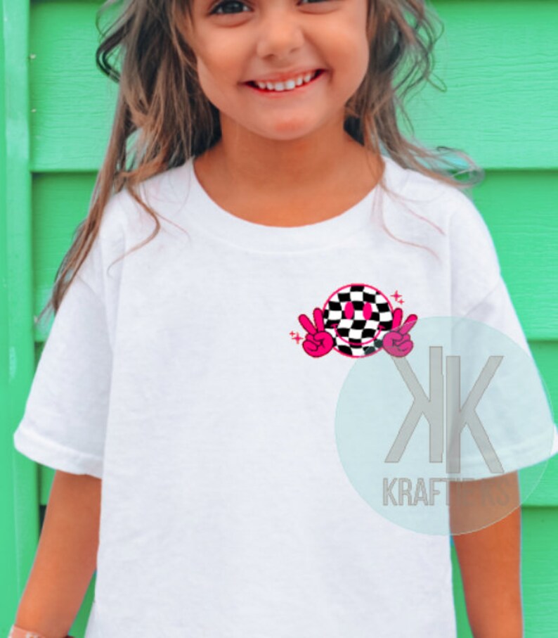 Puede incluir: Camiseta blanca con una cara sonriente a cuadros en negro y rosa con signos de paz. El gr&aacute;fico tiene dos estrellas rosas arriba.