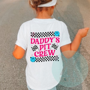 Puede incluir: Camiseta blanca con fondo a cuadros blanco y negro. El texto "Daddy's Pit Crew" est&aacute; en letras rosas con caritas sonrientes azules y banderas a cuadros a cada lado.