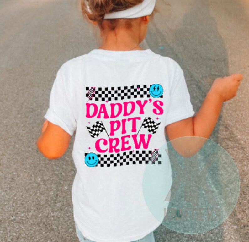 Puede incluir: Camiseta blanca con fondo a cuadros blanco y negro. El texto "Daddy's Pit Crew" est&aacute; en letras rosas con caritas sonrientes azules y banderas a cuadros a cada lado.