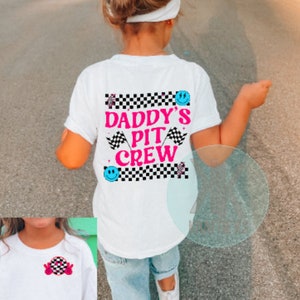 Puede incluir: Camiseta blanca con un dise&ntilde;o a cuadros blanco y negro y el texto "Daddy's Pit Crew" en letras rosas. El dise&ntilde;o tambi&eacute;n incluye una cara sonriente azul y una bandera de carreras rosa.