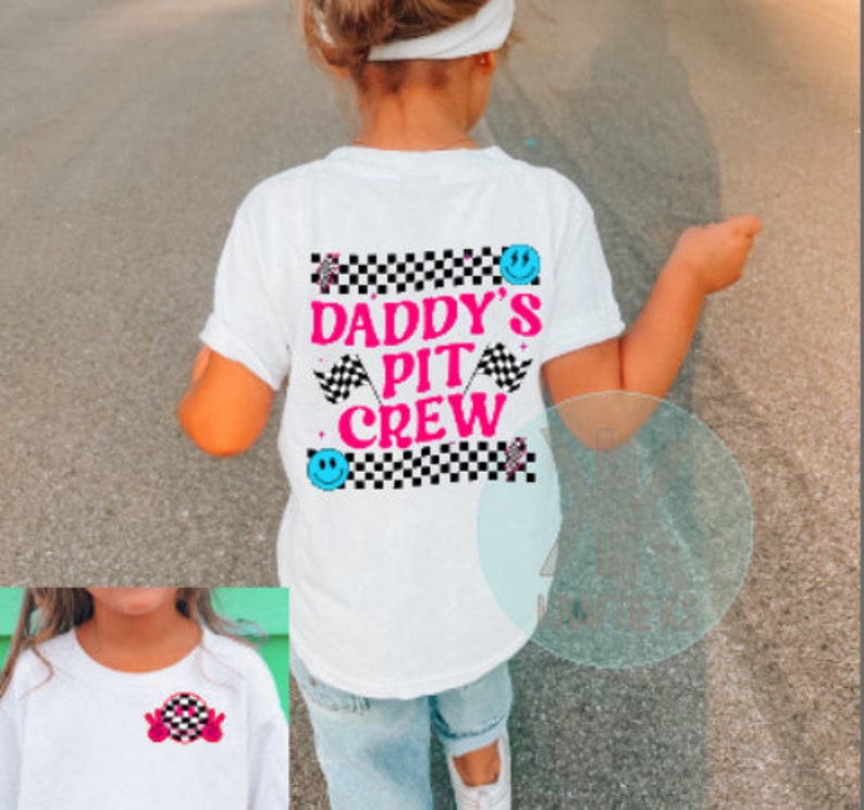 Puede incluir: Camiseta blanca con un dise&ntilde;o a cuadros blanco y negro y el texto "Daddy's Pit Crew" en letras rosas. El dise&ntilde;o tambi&eacute;n incluye una cara sonriente azul y una bandera de carreras rosa.
