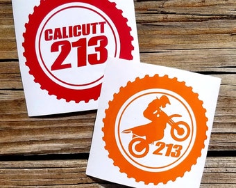 May include: Two white stickers with red and orange circular designs. The red sticker has the text "CALICUTT 213" in white. The orange sticker has a silhouette of a person riding a dirt bike with the number "213" below.