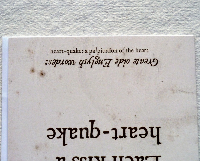 May include: A close-up of a vintage-style paper with the words "heart-quake" and "Greate oide Englysh wordes" printed in dark brown. The text "heart-quake: a palpitation of the heart" is also visible. The paper has a distressed, aged appearance.