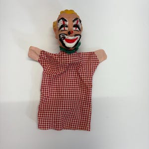 May include: A vintage clown hand puppet with a painted face, yellow hair, and a red and white checkered shirt. The puppet has a green collar and pink fabric arms. The clown's face features exaggerated red lips and black eye makeup.
