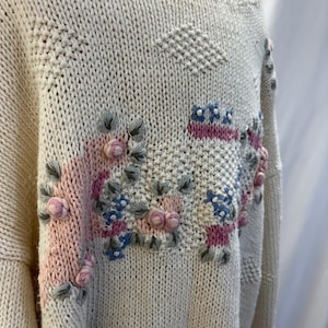 May include: Cream-colored knit sweater with floral embroidery. The sweater features a crew neck and long sleeves. The embroidery includes pink, blue, and gray flowers and leaves, adding a decorative touch to the garment.
