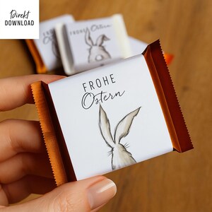 May include: A white chocolate bar wrapper with the words "FROHE OSTERN" and a drawing of a bunny. The wrapper has a brown border and is held in a hand. The background is a blurred wooden surface.