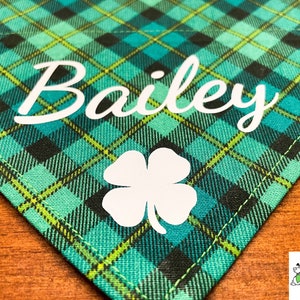 May include: Green and black plaid fabric dog bandana with the name "Bailey" and a white four-leaf clover.