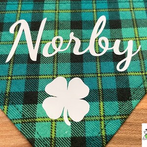 May include: A green and black plaid dog bandana with the name "Norby" and a white four-leaf clover.