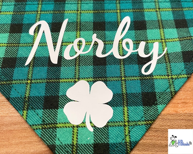 May include: A green and black plaid dog bandana with the name "Norby" and a white four-leaf clover.