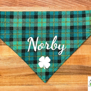 May include: Green and black plaid dog bandana with a white shamrock and the name "Norby" written in white cursive.