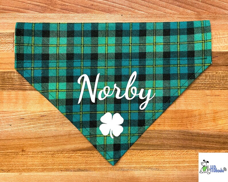 May include: Green and black plaid dog bandana with a white shamrock and the name "Norby" written in white cursive.