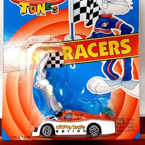 Puede incluir: Un coche de juguete de fundición Matchbox Looney Tunes Racers con Bugs Bunny en un coche de carreras blanco y naranja con una bandera a cuadros. El embalaje es azul y naranja con el logotipo de Looney Tunes y las palabras "Racers" en letras grandes.