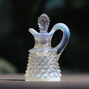 May include: A clear glass creamer with a handle and a textured, hobnail-like pattern. The creamer has a decorative stopper and a light blue iridescent sheen. The creamer is approximately 4 inches tall.