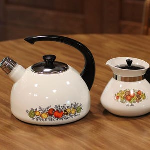 May include: A white enamel tea kettle and a matching creamer pitcher, both featuring a colorful vegetable and fruit pattern. The kettle has a black handle, a black lid knob, and a silver spout. The creamer has a black handle and a black lid.