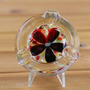 May include: A clear glass ashtray with a red, black, and white flower design. The ashtray is on a clear plastic stand.