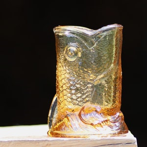 May include: A decorative glass fish-shaped vase in amber color. The vase has a detailed scale pattern and fin details. The fish's mouth forms the opening of the vase. The vase is sitting on a light-colored surface against a black background.