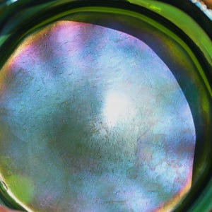 May include: Close-up of a green glass bowl with iridescent interior. The bowl's surface reflects a spectrum of colors, including blue, purple, and yellow. Gold floral designs adorn the rim, adding a decorative touch to the vintage glassware.
