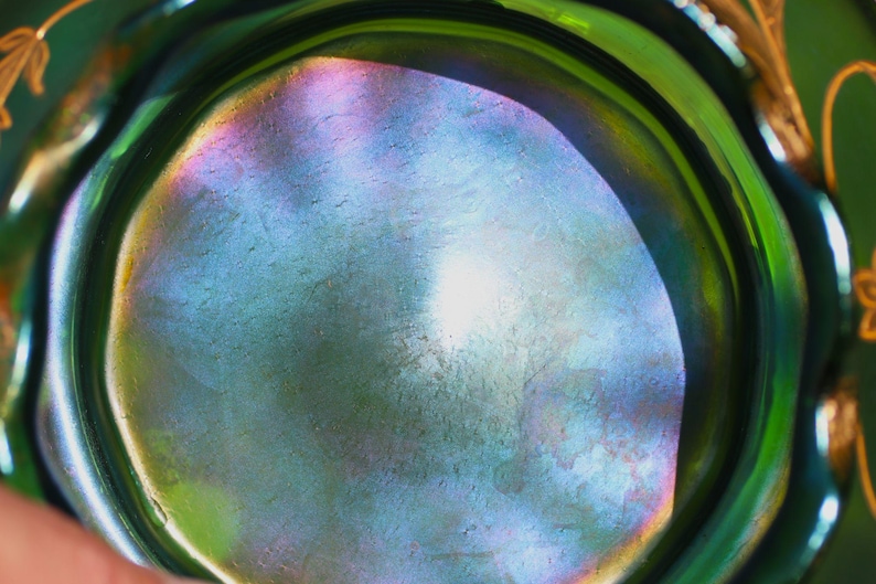 May include: Close-up of a green glass bowl with iridescent interior. The bowl's surface reflects a spectrum of colors, including blue, purple, and yellow. Gold floral designs adorn the rim, adding a decorative touch to the vintage glassware.