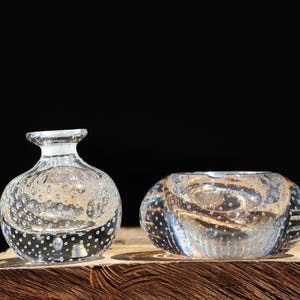 May include: Two clear glass bubble vases, one round and one bowl-shaped, sit on a wooden surface. The vases have a textured, bubbly surface.