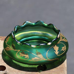 May include: A green iridescent glass bowl with a ruffled rim and gold floral designs. The bowl has a round base and a wide opening. The gold floral designs are on the side of the bowl.