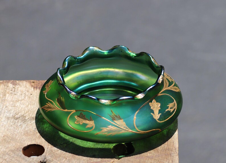 May include: A green iridescent glass bowl with a ruffled rim and gold floral designs. The bowl has a round base and a wide opening. The gold floral designs are on the side of the bowl.