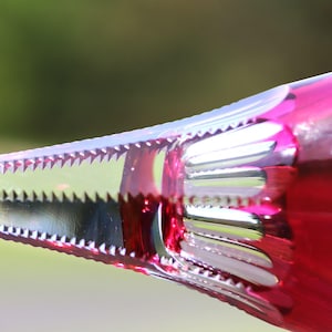 May include: A close-up view of a vibrant magenta glass object featuring intricate, sharp-edged geometric cuts and a serrated, saw-tooth pattern along the stem. The glass displays a deep, translucent pink hue with polished, reflective facets.