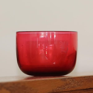 May include: A vibrant, ruby-red glass bowl with a rounded shape. The bowl sits on a wooden surface, with a light-colored background. The glass appears to be transparent, with a slight gradient in color from top to bottom.