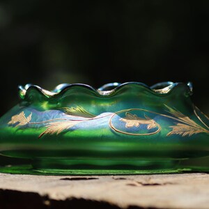 May include: A green glass bowl with a ruffled rim and gold floral designs. The bowl has a wide, shallow shape and sits on a small base. The glass has an iridescent sheen, reflecting light.