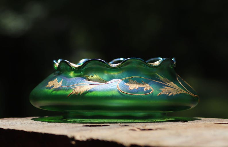 May include: A green glass bowl with a ruffled rim and gold floral designs. The bowl has a wide, shallow shape and sits on a small base. The glass has an iridescent sheen, reflecting light.