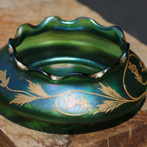 May include: A green iridescent glass bowl with a ruffled rim and gold floral designs. The bowl has a wide, shallow shape and sits on a wooden surface. The floral design is painted in gold.
