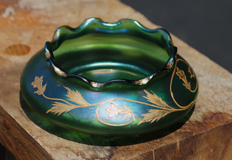 May include: A green iridescent glass bowl with a ruffled rim and gold floral designs. The bowl has a wide, shallow shape and sits on a wooden surface. The floral design is painted in gold.