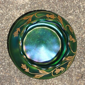 May include: A green iridescent glass bowl with a scalloped rim and gold leaf design. The bowl has a circular shape and a dark green exterior with a shimmering interior. The gold leaf design is a repeating pattern around the rim.