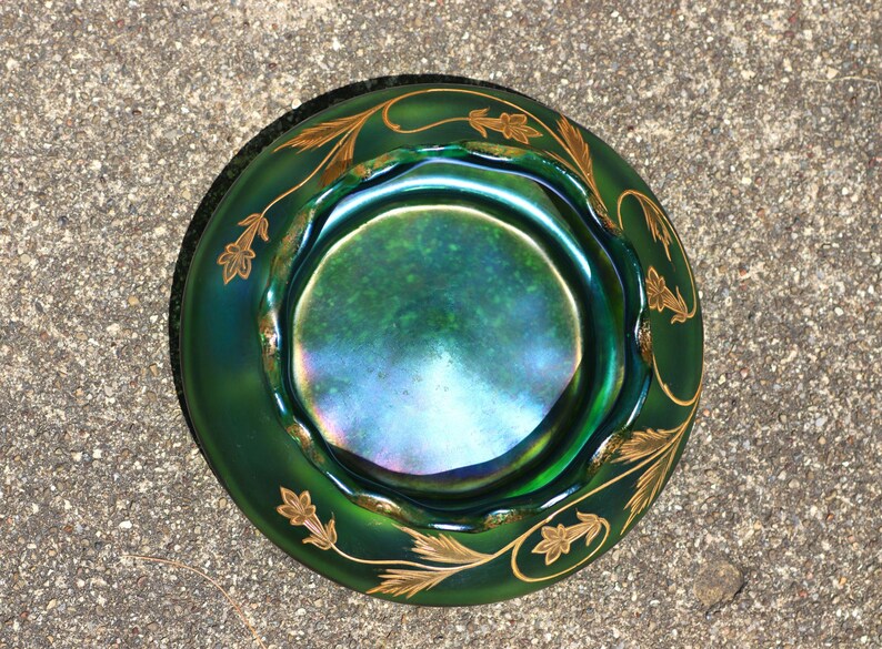 May include: A green iridescent glass bowl with a scalloped rim and gold leaf design. The bowl has a circular shape and a dark green exterior with a shimmering interior. The gold leaf design is a repeating pattern around the rim.