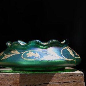 May include: A green iridescent glass bowl with a ruffled rim and gold leaf design. The bowl has a wide, shallow shape and sits on a small base. The gold leaf design features stylized floral and leaf patterns.