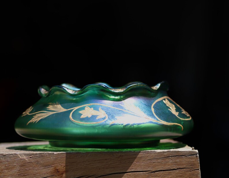 May include: A green iridescent glass bowl with a ruffled rim and gold leaf design. The bowl has a wide, shallow shape and sits on a small base. The gold leaf design features stylized floral and leaf patterns.