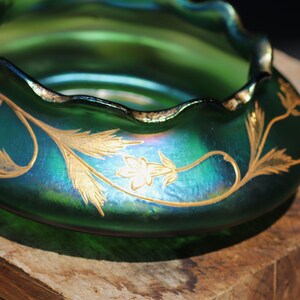 May include: A green iridescent glass bowl with a ruffled rim and gold floral designs. The bowl has a rounded shape and sits on a wooden surface. The gold floral pattern is a repeating design around the bowl.