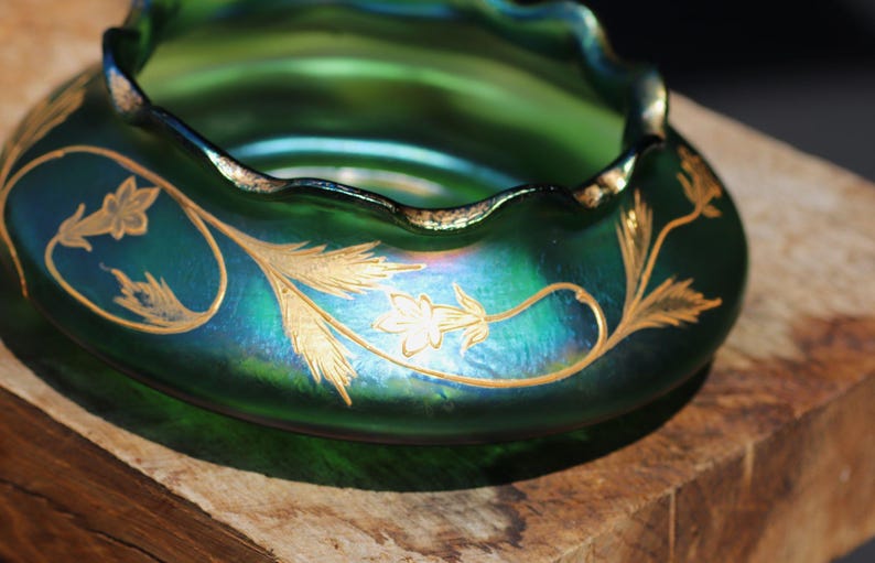 May include: A green iridescent glass bowl with a ruffled rim and gold floral designs. The bowl has a rounded shape and sits on a wooden surface. The gold floral pattern is a repeating design around the bowl.
