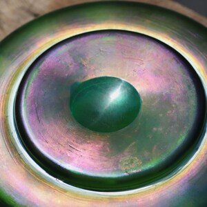 May include: A close-up of a decorative glass bowl with a green and iridescent finish. The bowl has a central, raised, rounded feature. The colors shift from green to purple and gold.
