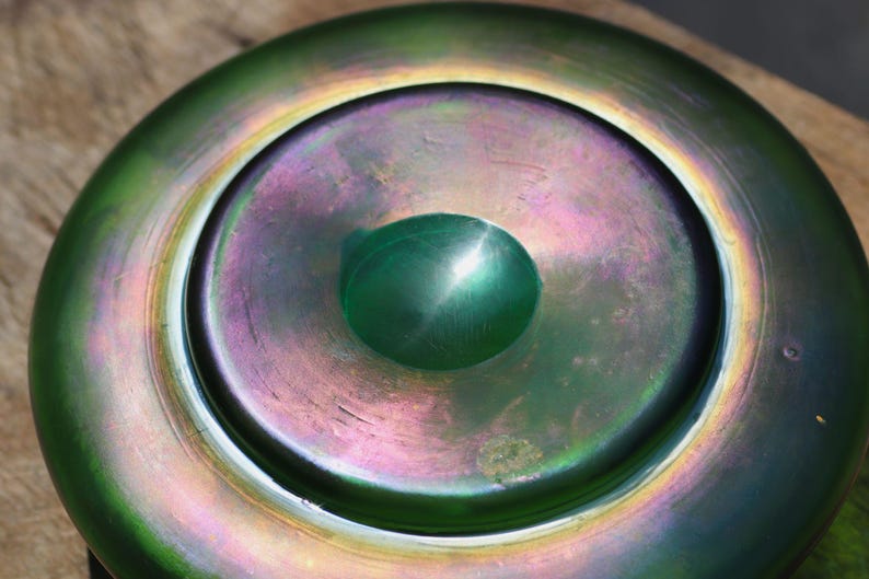 May include: A close-up of a decorative glass bowl with a green and iridescent finish. The bowl has a central, raised, rounded feature. The colors shift from green to purple and gold.