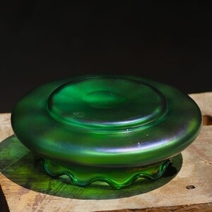 May include: A green iridescent glass bowl with a wide, shallow design. The bowl has a circular base and a slightly raised center. The rim has a wavy, scalloped edge. The glass reflects a range of colors, including green and purple.