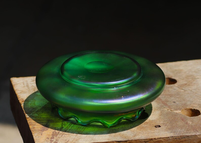 May include: A green iridescent glass bowl with a wide, shallow design. The bowl has a circular base and a slightly raised center. The rim has a wavy, scalloped edge. The glass reflects a range of colors, including green and purple.