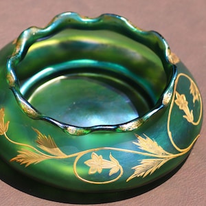 May include: A green iridescent glass bowl with a ruffled rim and gold floral designs. The bowl has a rounded shape and a glossy finish. The gold floral designs are painted on the exterior of the bowl.